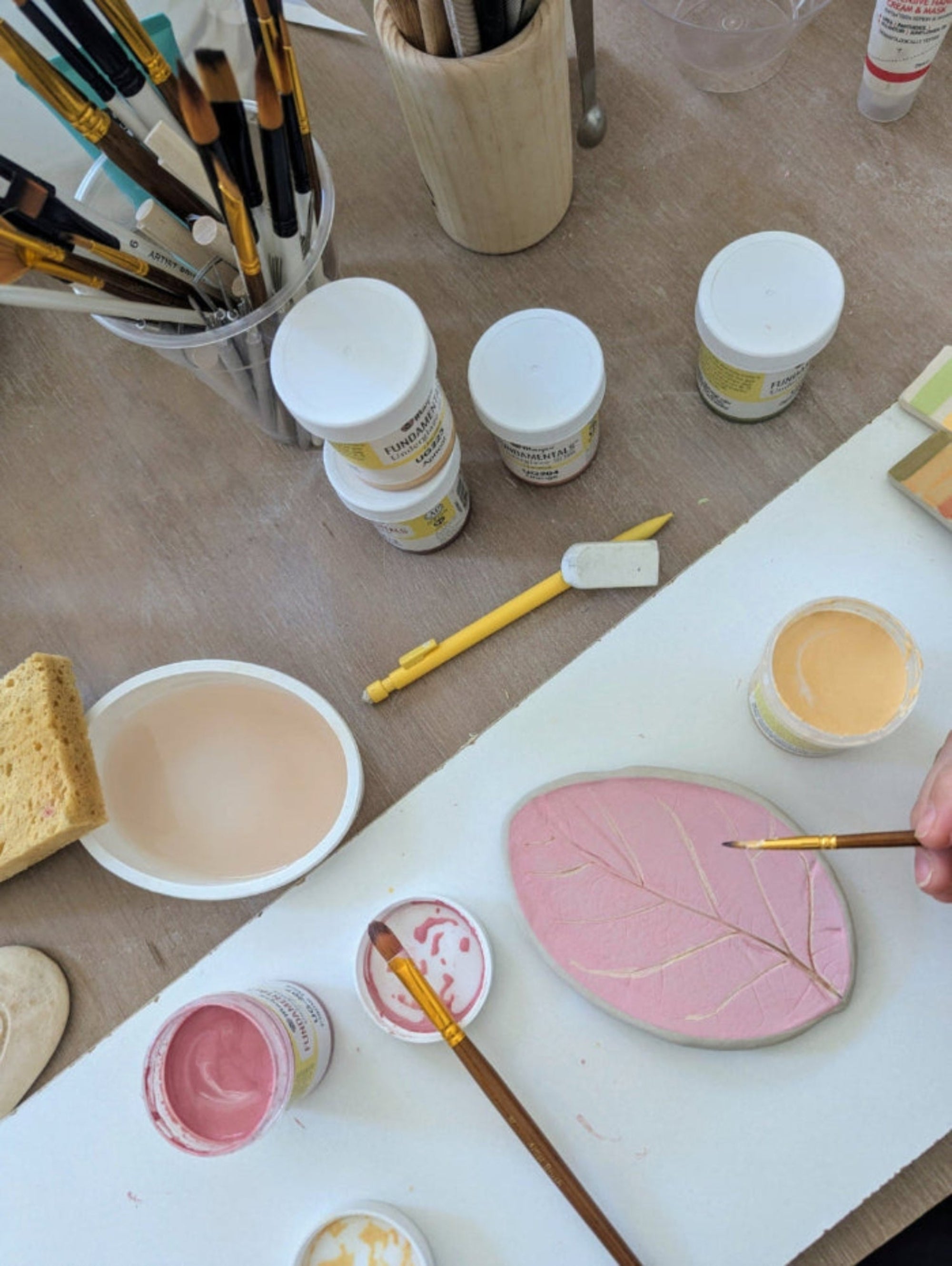 Artistic workspace with paint containers, brushes, and a red ceramic piece in the shape of a leaf being painted.